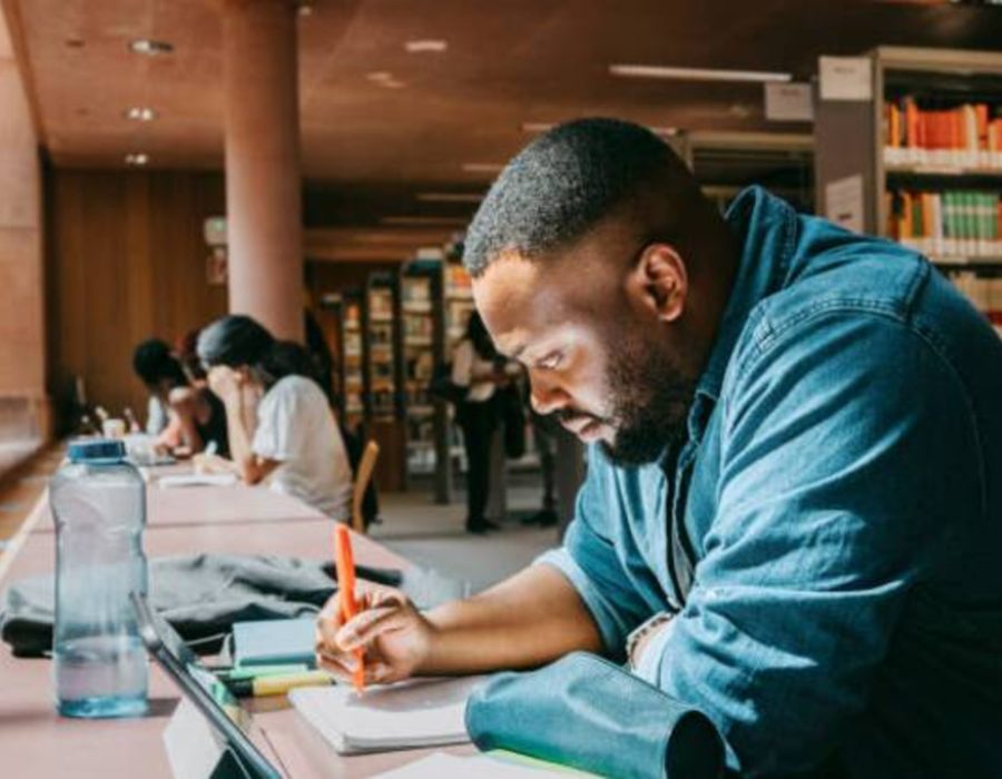 https-media-gettyimages-com-id-1439945611-photo-student-writing-in-notebook-while-sitting-with-tablet-pc-in-college-library (1)